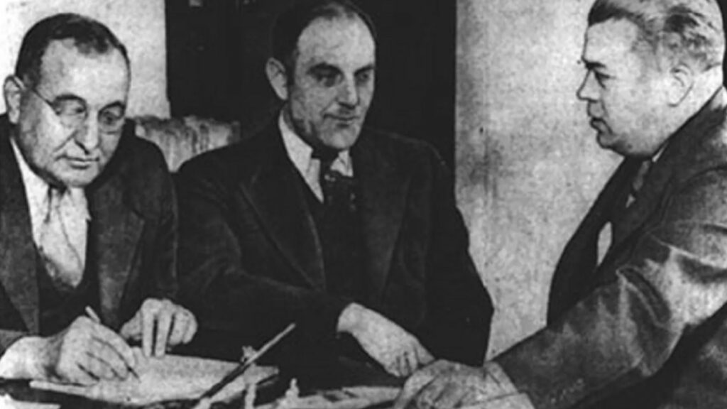 Viktor Lustig (uprostřed) při policejním výslechu v roce 1935. Zdroj: Chicago Bureau (Federal Bureau of Investigation) - Wide World Photos. – Item Catalog Cotta Have It! Collectibles, Inc., Volné dílo, https://commons.wikimedia.org/w/index.php?curid=35462819